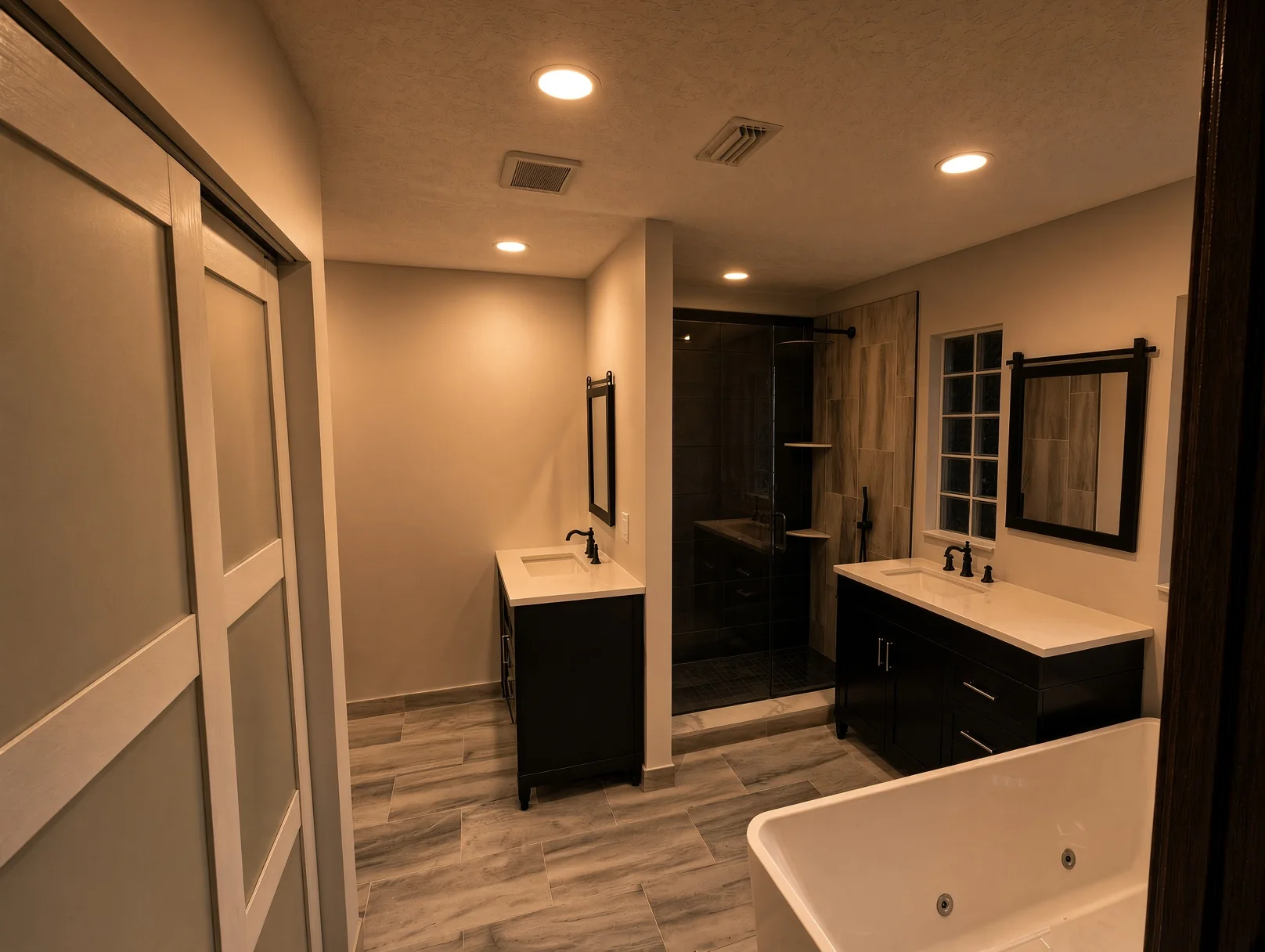 Bathroom with two black vanities, a dark tile shower and a soaking tub in warm light