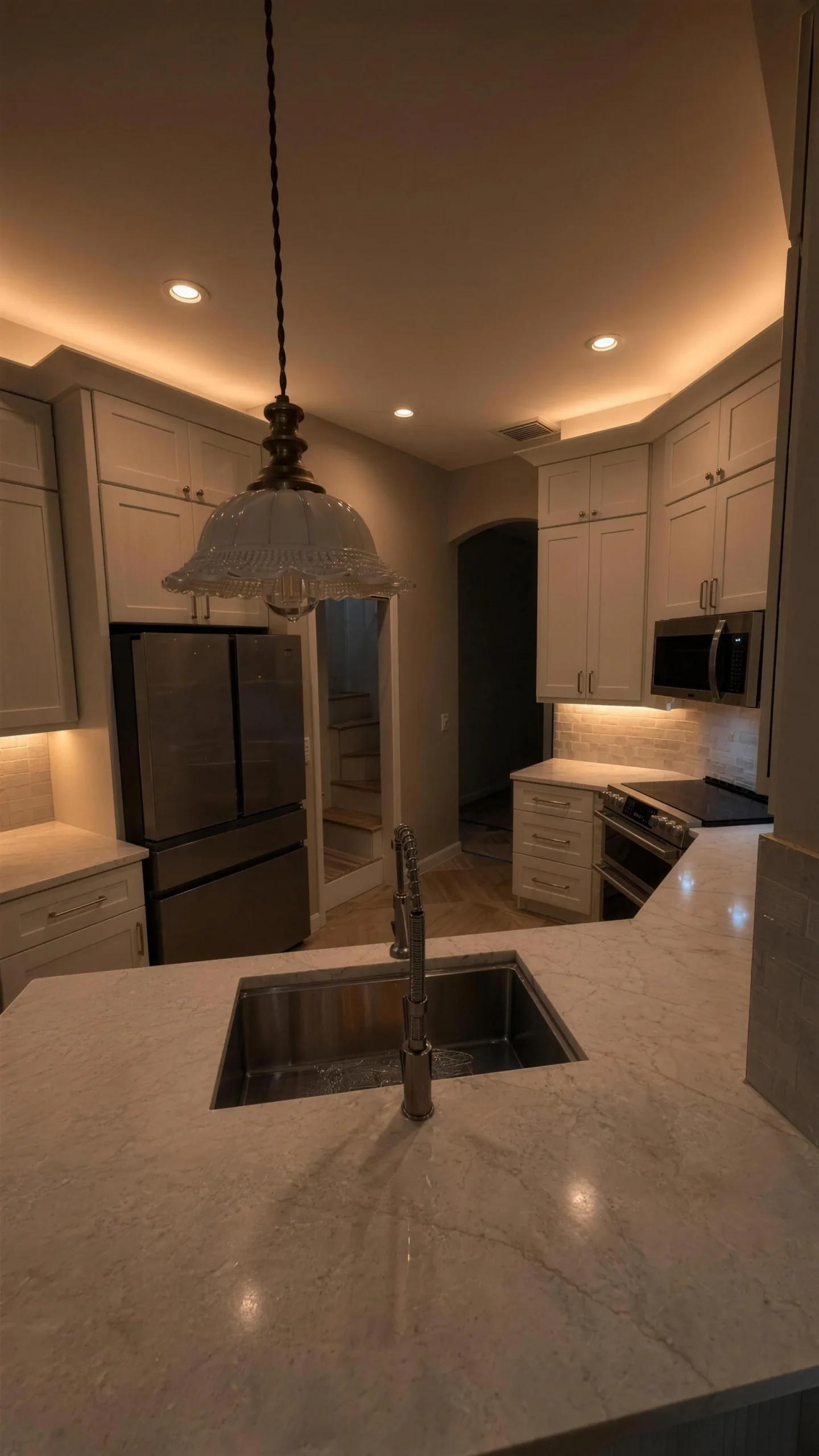 Kitchen at night with a crystal pendant over a marble island and white cabinetry