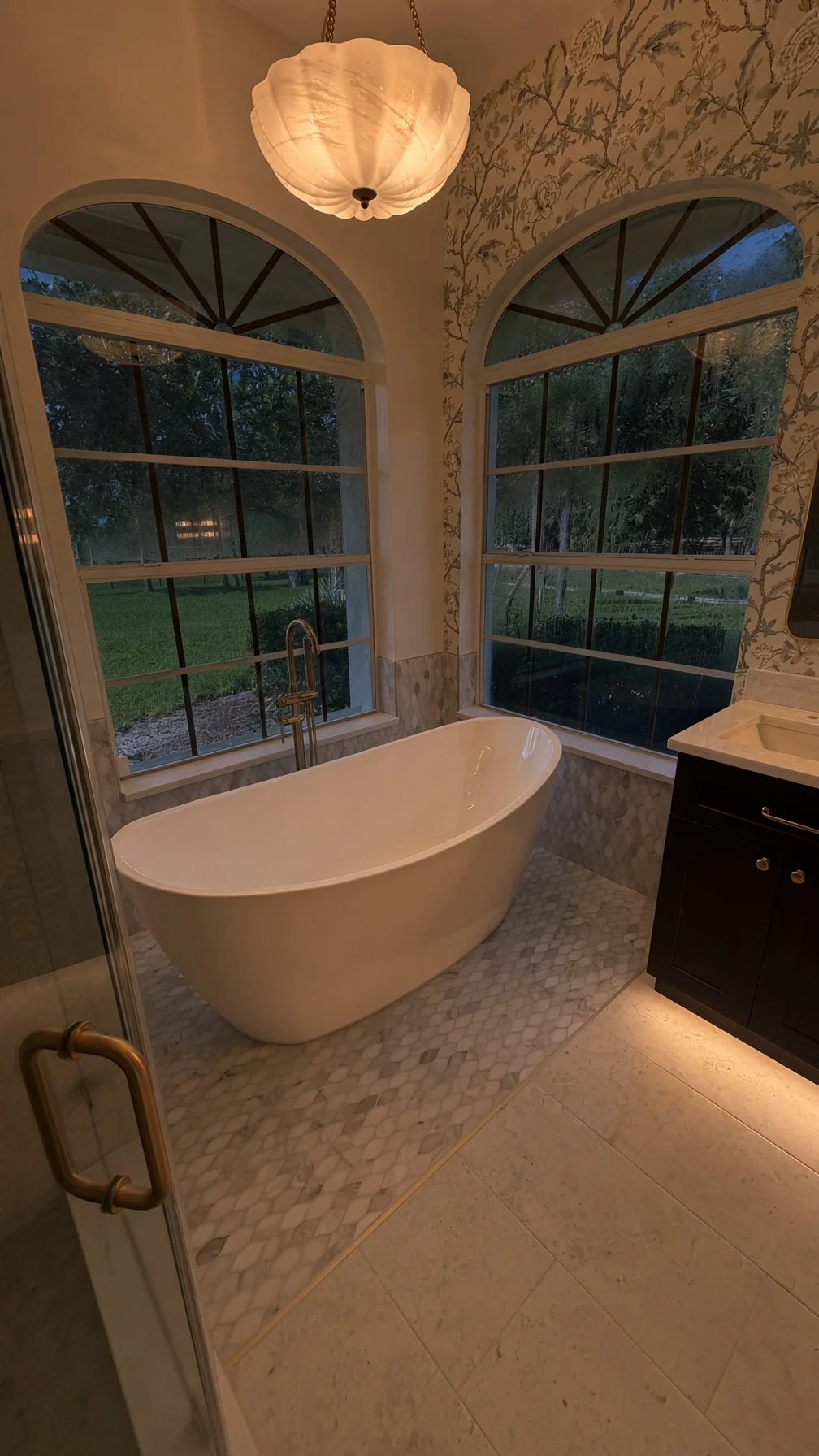 Freestanding soaking tub between two arched windows at dusk under a shell pendant