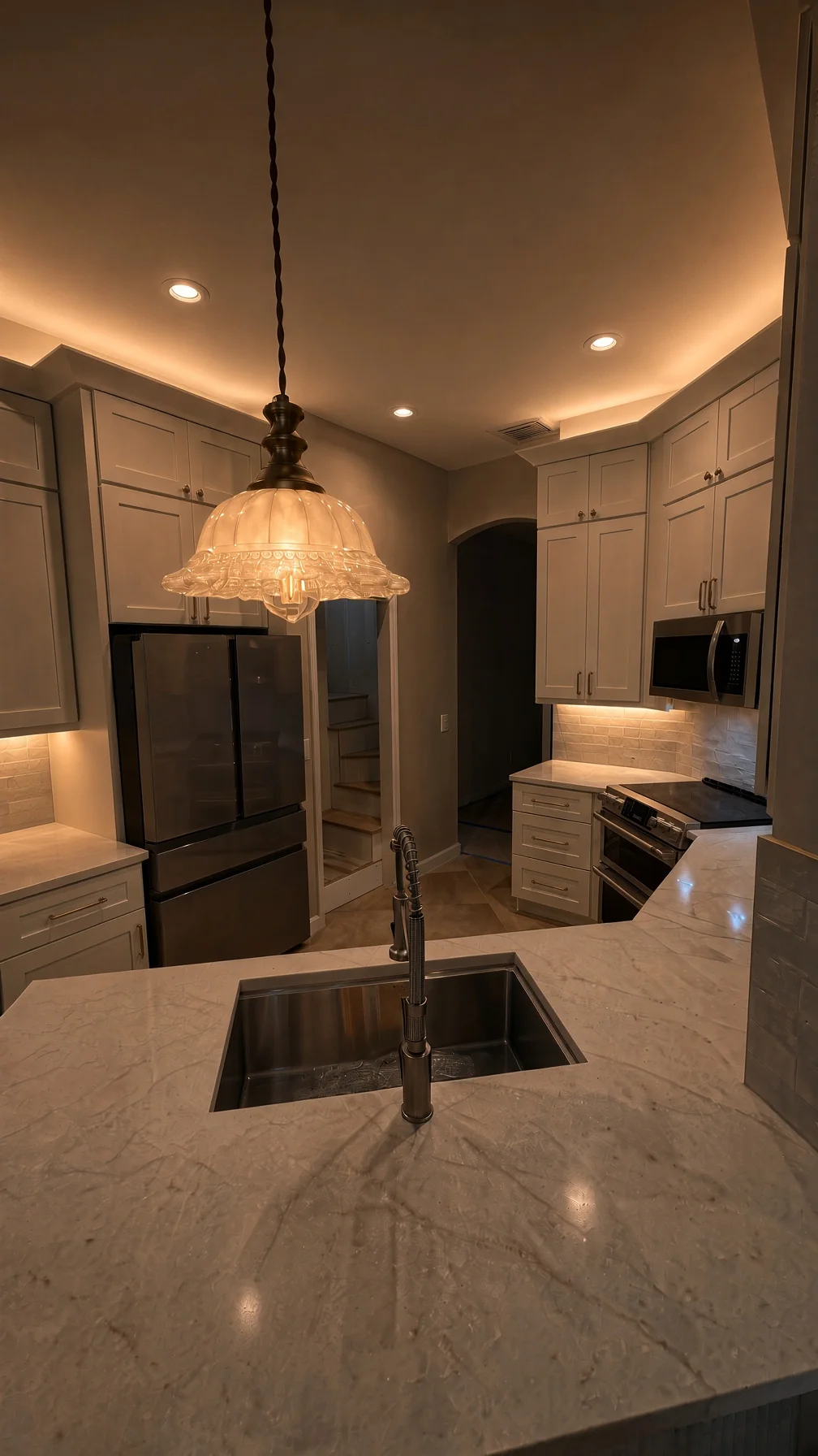 Kitchen at night with a lit crystal pendant over a marble island and white cabinetry