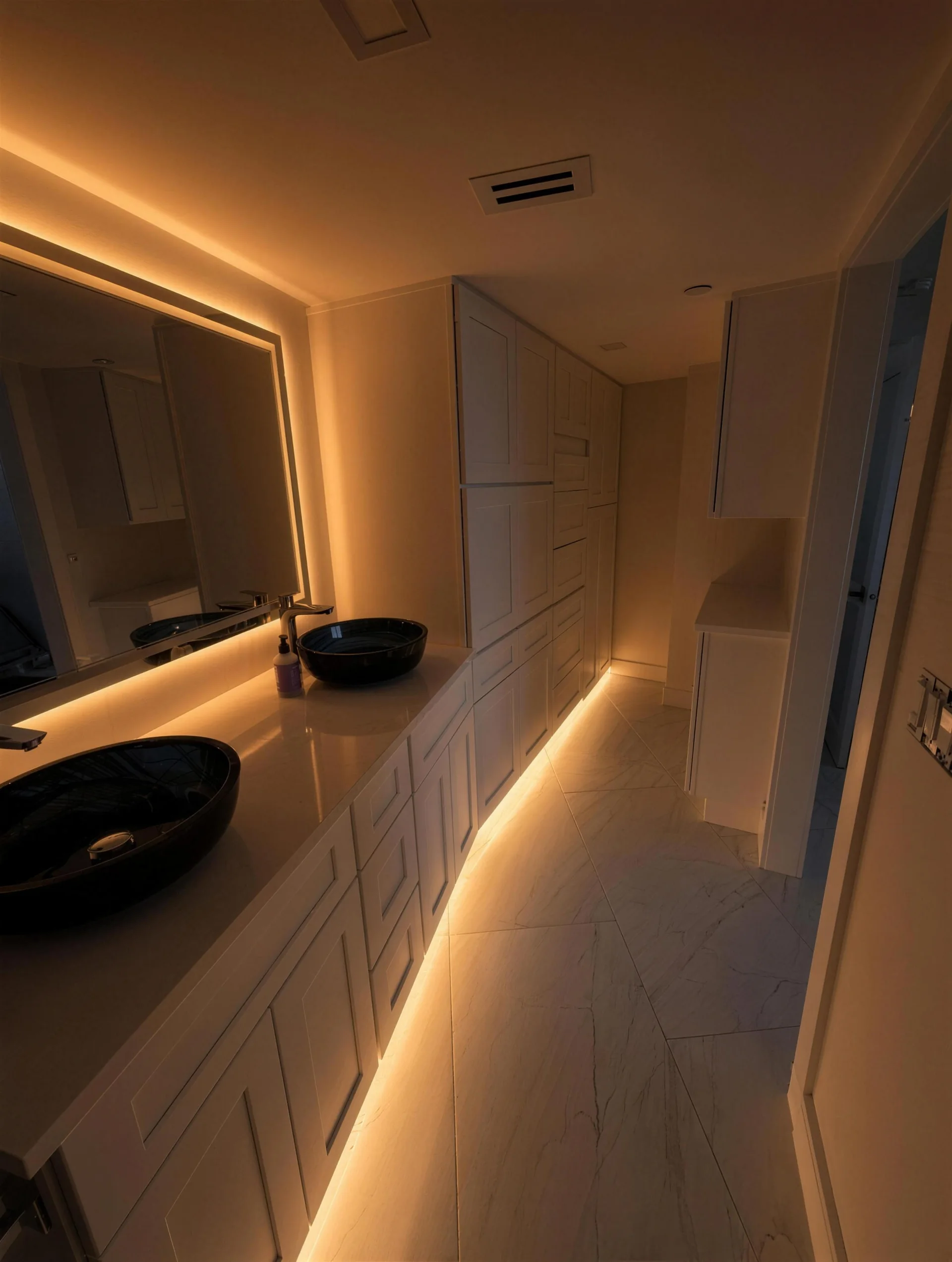 Bathroom vanity at night with two vessel sinks, a lit mirror and glowing under-cabinet lighting