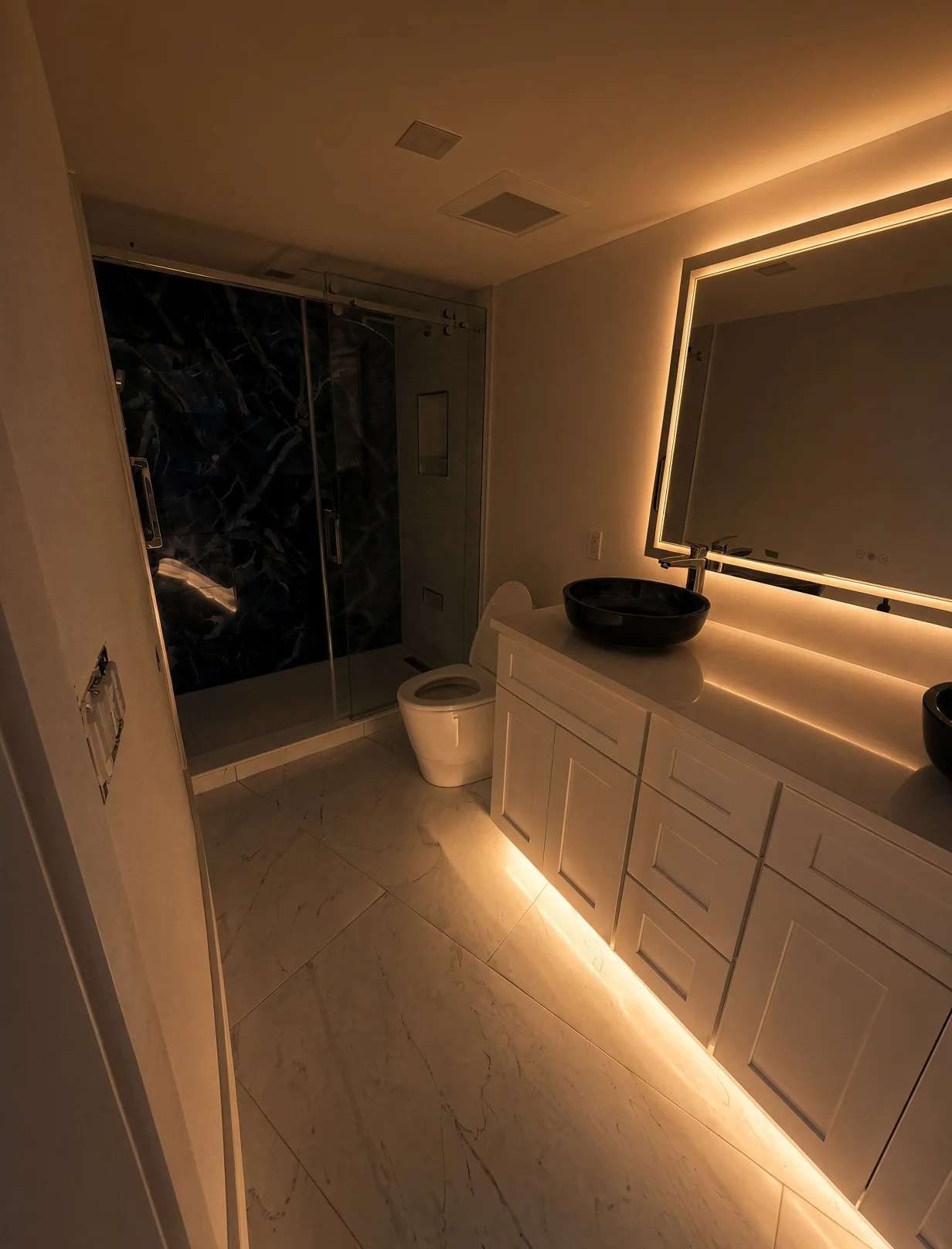 Dark marble glass shower and white vanity lit by an LED mirror and toe-kick lighting
