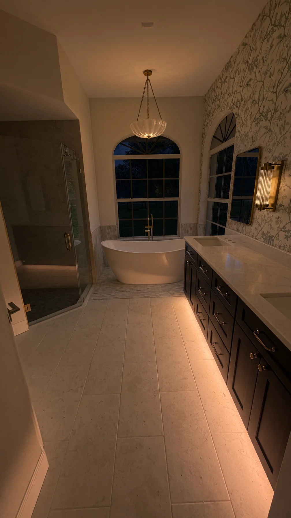 Long bathroom at night with a lit dark vanity, freestanding tub and arched window