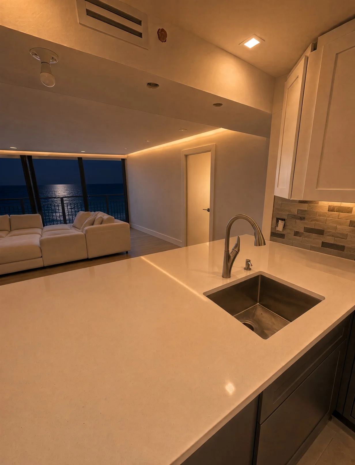 Kitchen at night with a quartz counter looking across a living room to the moonlit ocean